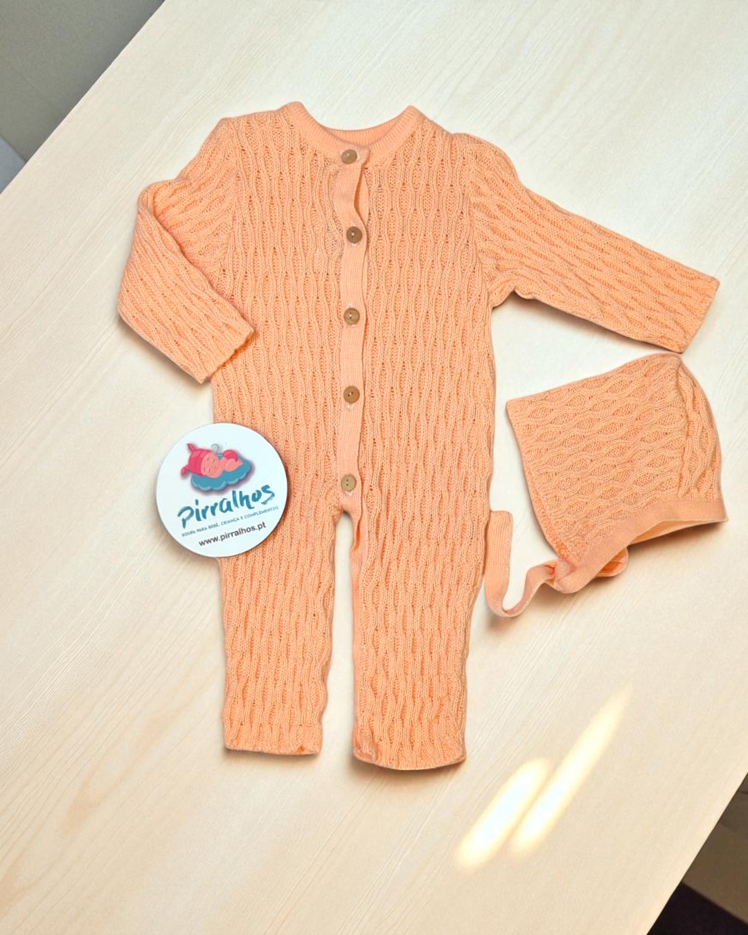Macacão de bebé cor de pêssego com padrões trançados e botões de madeira, com touca igual e etiqueta PIRRALHOS sobre mesa clara