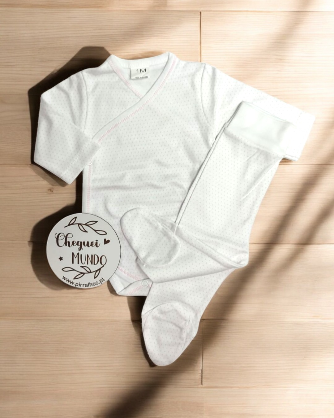 Roupa de bebé branca com pontos e calças com pés sobre chão de madeira clara, com círculo de papel ao lado