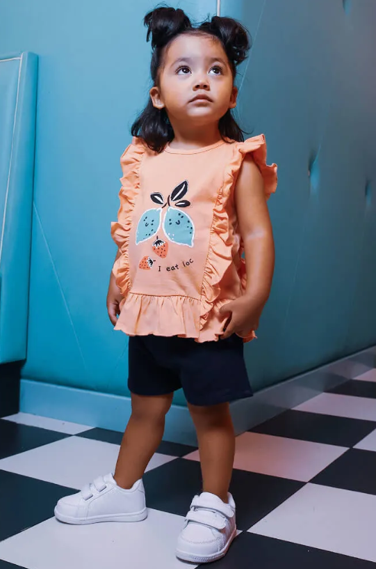 Criança com t-shirt cor de pêssego com estampa de pêssegos, calções pretos e ténis brancos num chão xadrez.