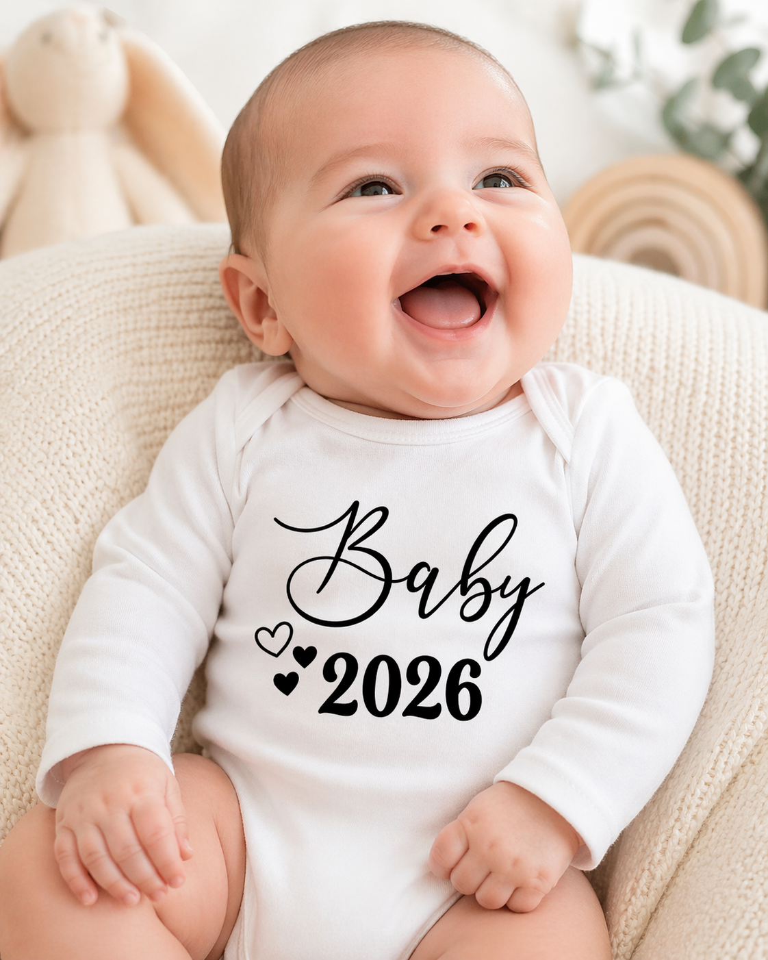 Bebé sorridente com body branco de manga comprida com texto Baby 2026 preto, sentado numa almofada creme com brinquedo e planta ao fundo.
