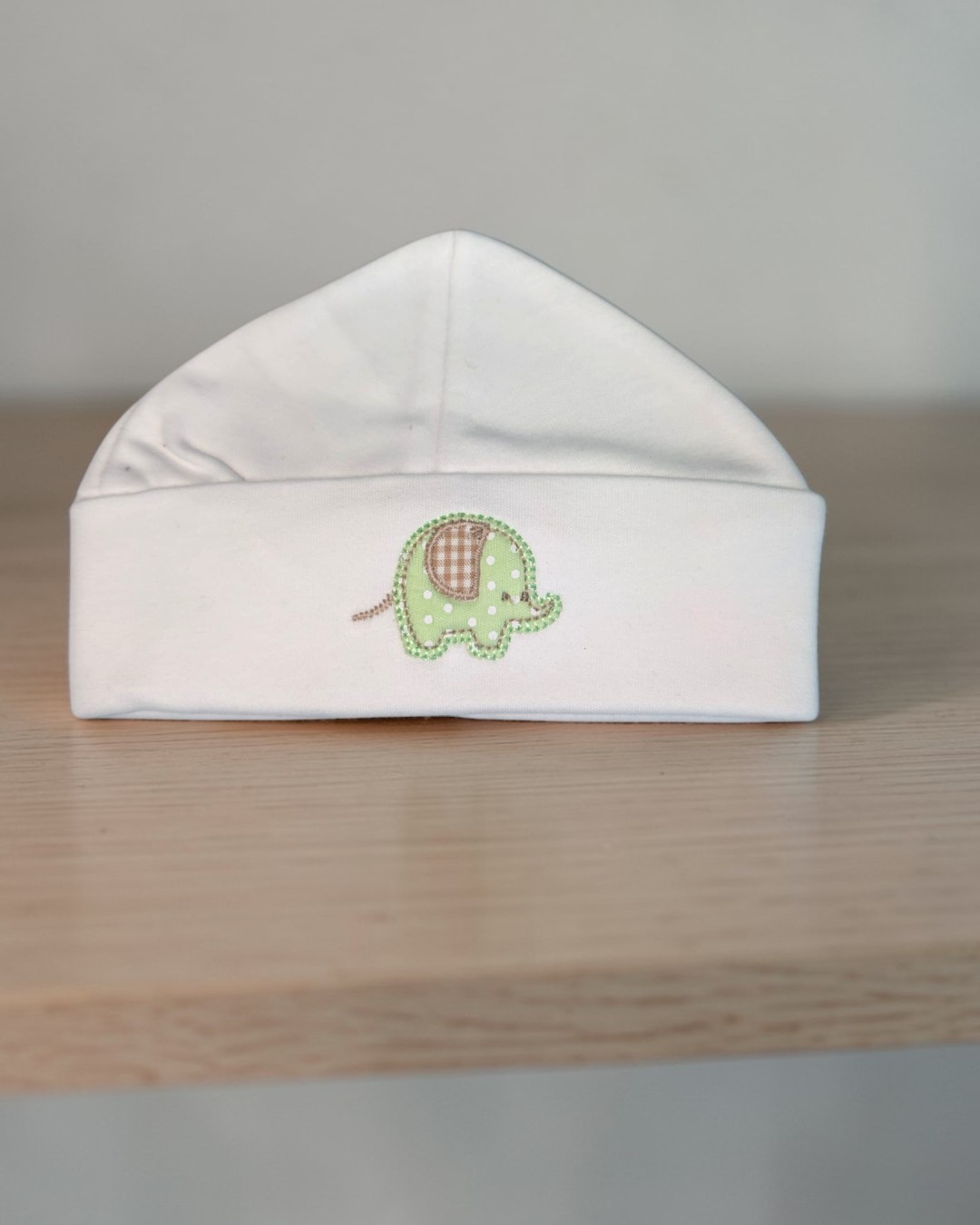 Gorro branco de bebé com bordado de elefante verde sobre superfície de madeira