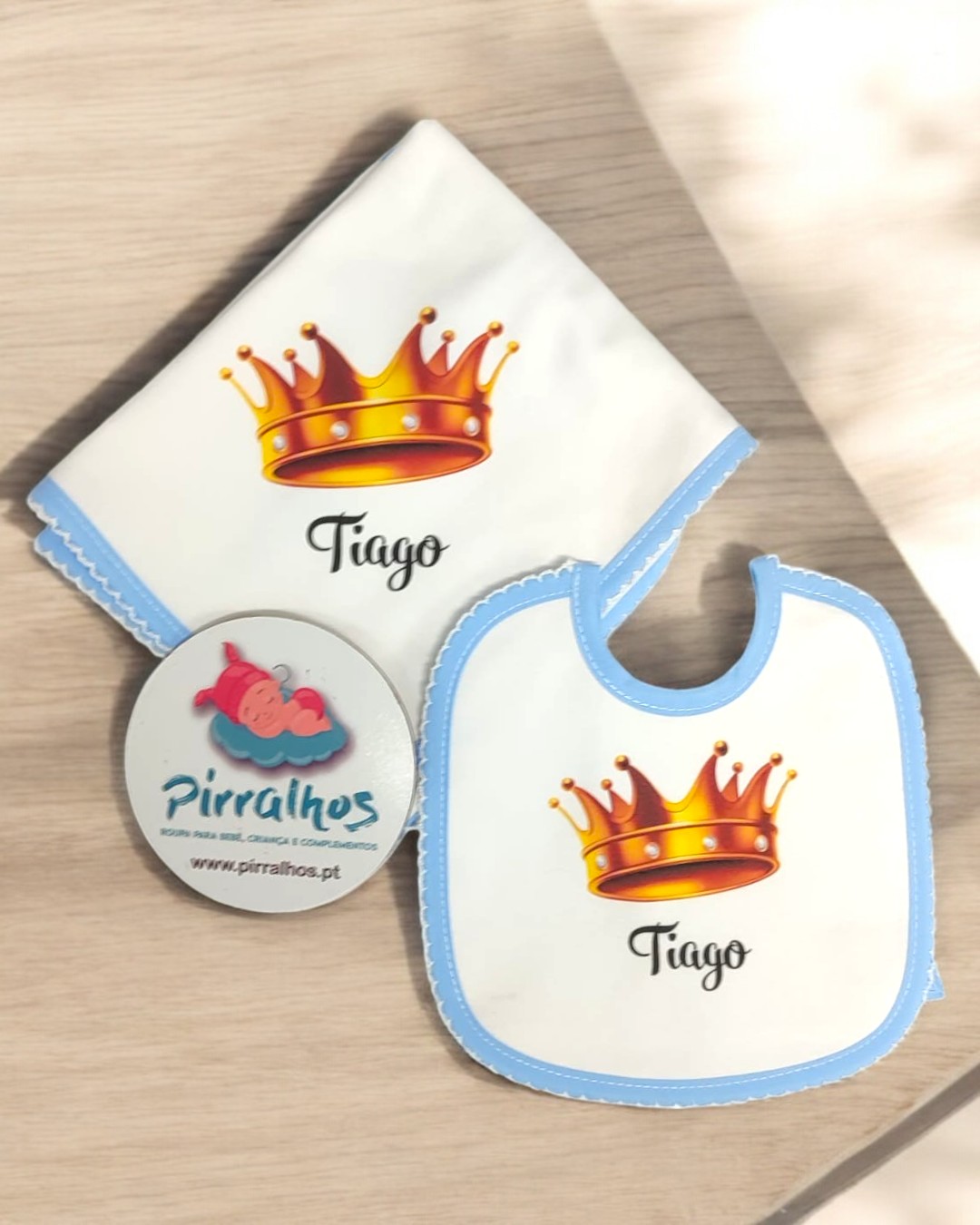 Conjunto de babete e fralda brancos com bordas azuis, coroa laranja e nome Tiago sobre mesa de madeira