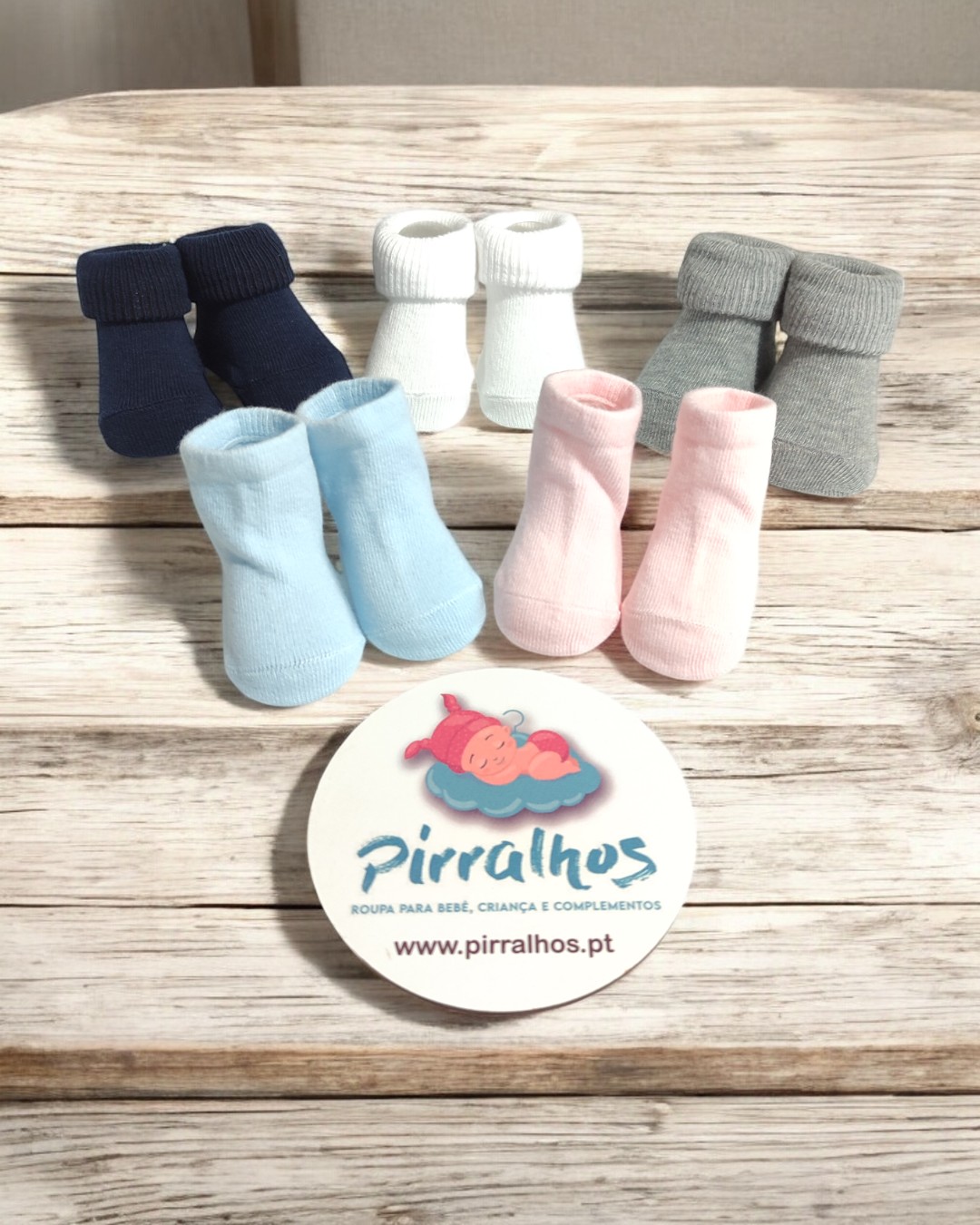 Cinco pares de meias de bebé em várias cores sobre madeira com cartão publicitário da Pirralhos