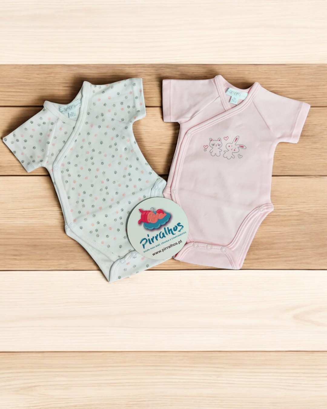Dois bodies de bebé, um branco com estrelas coloridas e outro rosa claro com bordado, sobre madeira clara com etiqueta redonda.
