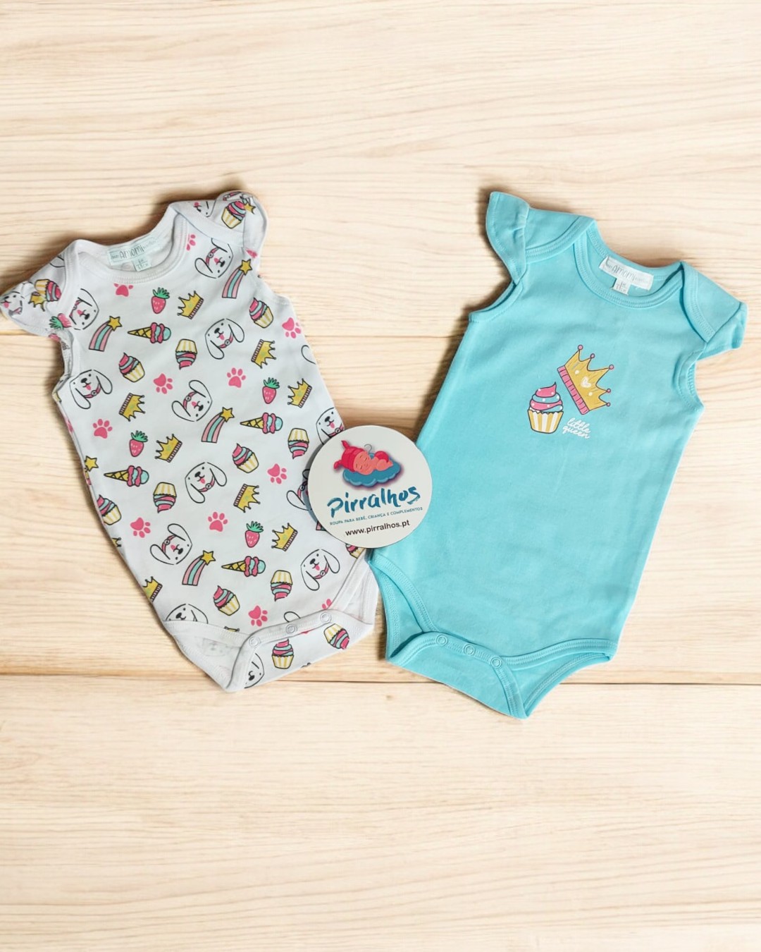 Dois bodies de bebé em algodão, um branco com padrão colorido e outro azul claro, sobre madeira clara