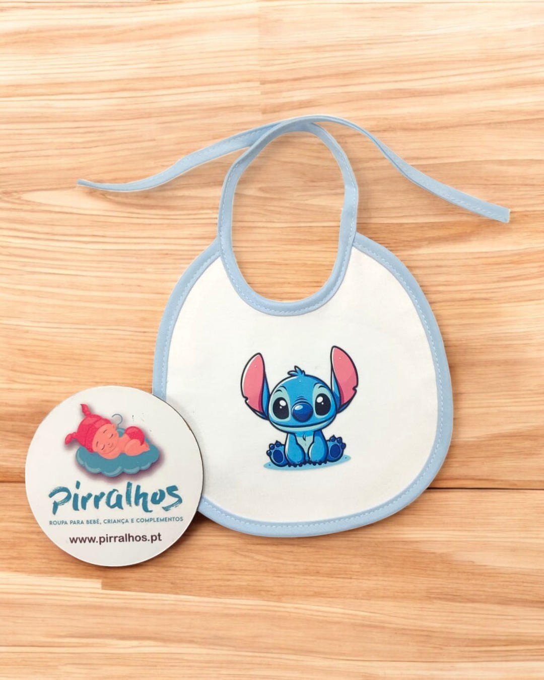 Babete branco com desenho azul do Stitch e selo redondo da marca Pirralhos em madeira clara
