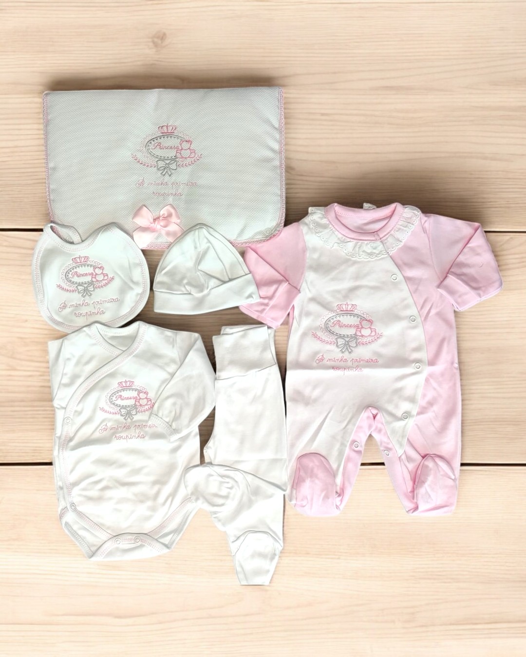 Conjunto de roupa de bebé rosa e branca com bordados, dispostos sobre superfície de madeira clara.