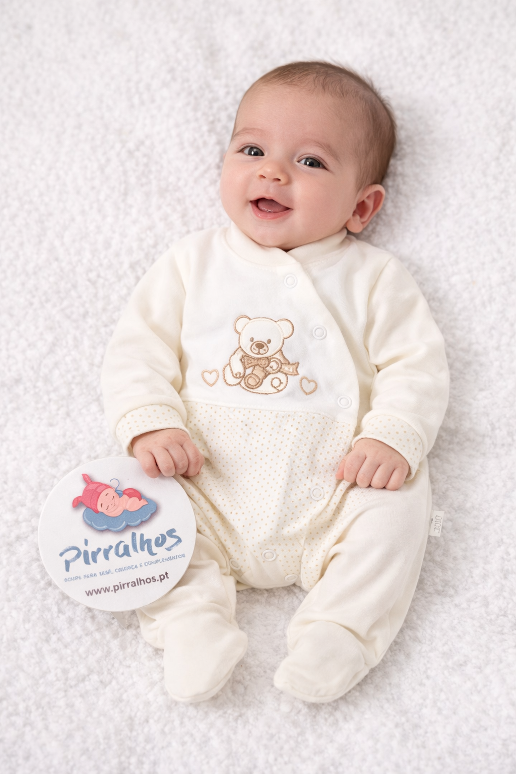 Bebé com macacão creme e estampado de ursinho, segurando etiqueta redonda com o texto PIRRALHOS