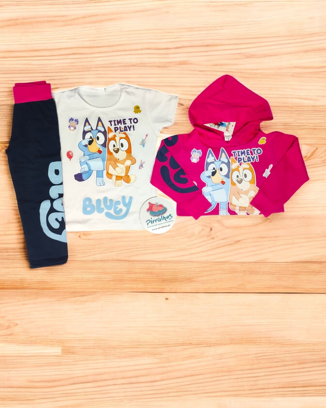 Conjunto infantil com camisola branca, calças azul escuro e casaco rosa com estampados de personagens e texto