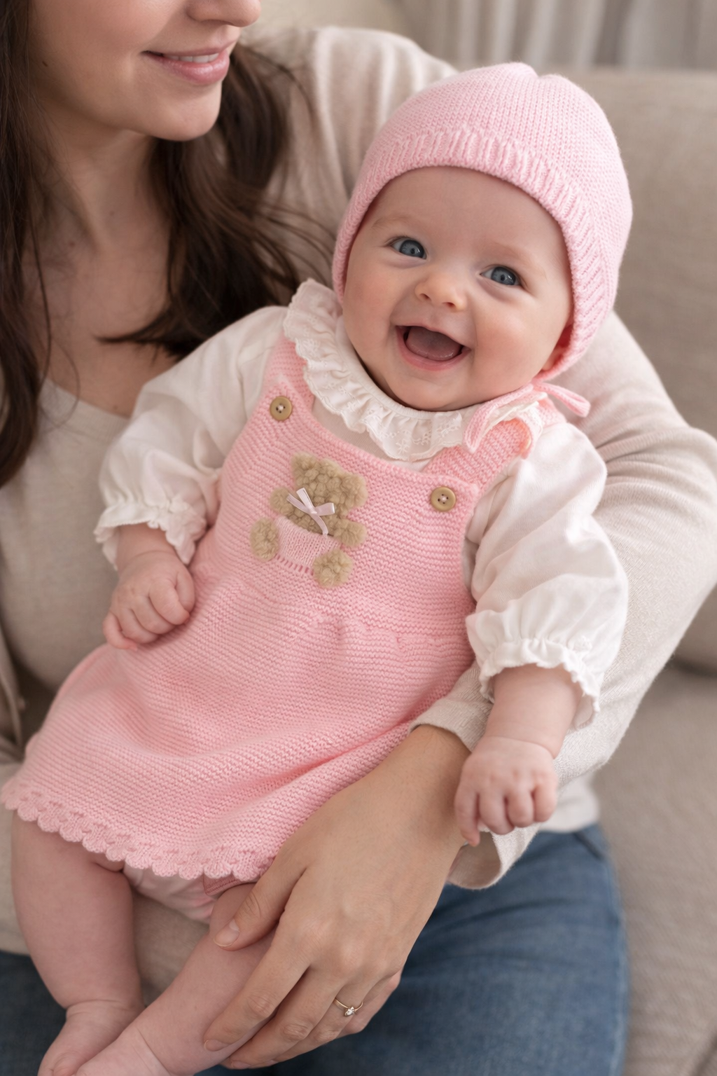 Bebé sorridente com vestido rosa de malha, camisola branca com folhos e gorro rosa, nos braços de uma mulher.