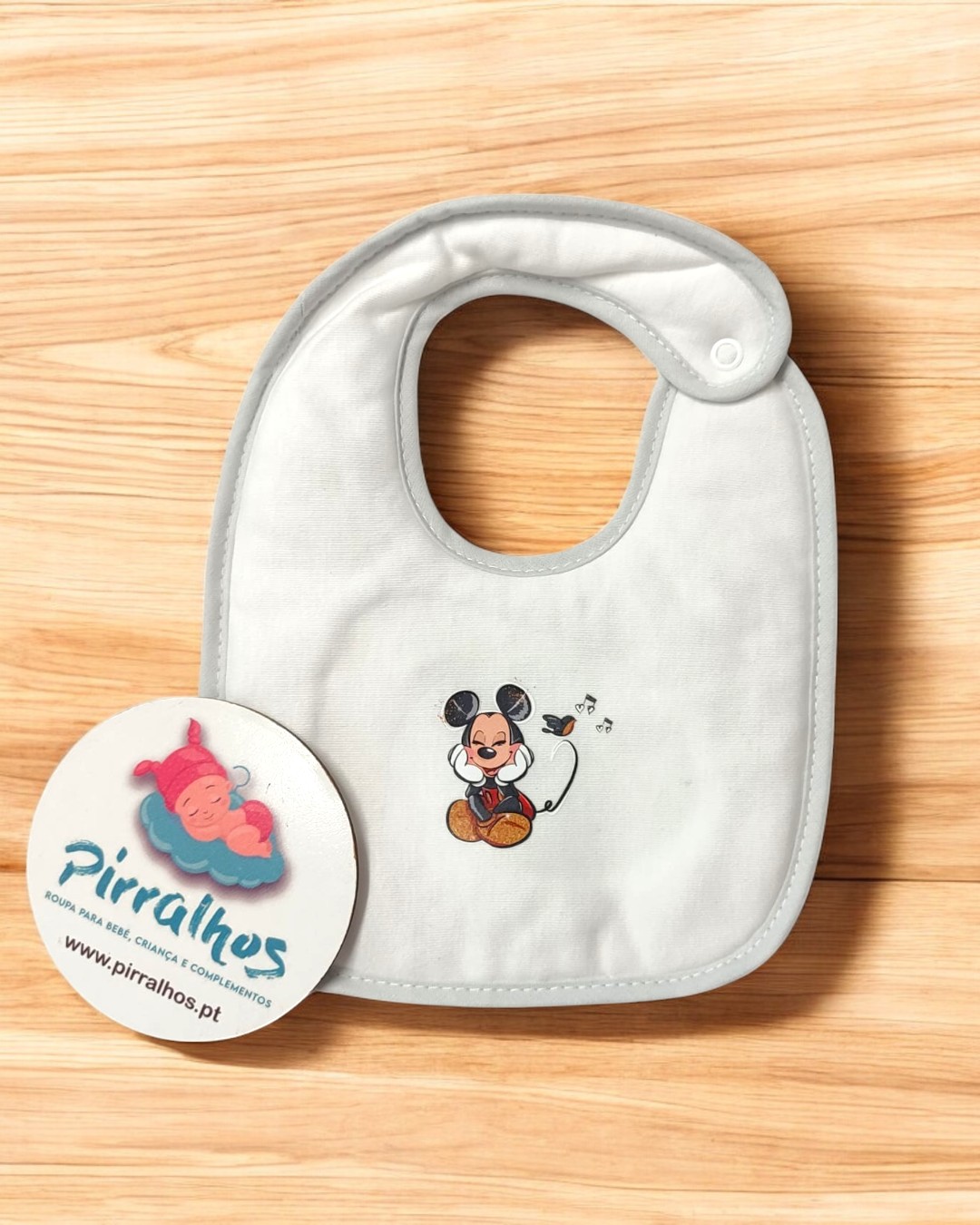 Babete para bebé branco com desenho do Mickey Mouse, sobre superfície de madeira clara com cartão promocional