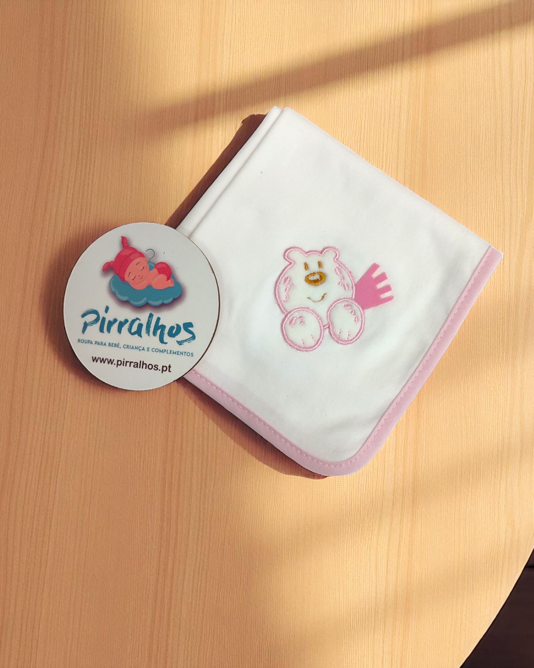 Fralda branca com borda rosa e desenho de ursinho, etiqueta Pirralhos no fundo de madeira