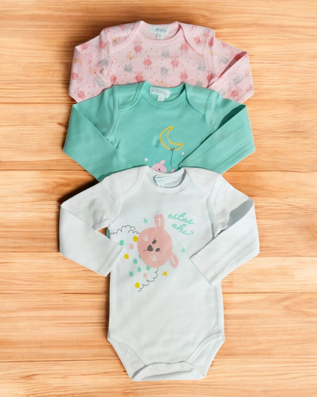 Três bodies de bebé com manga comprida em várias cores e padrões sobre fundo de madeira clara.