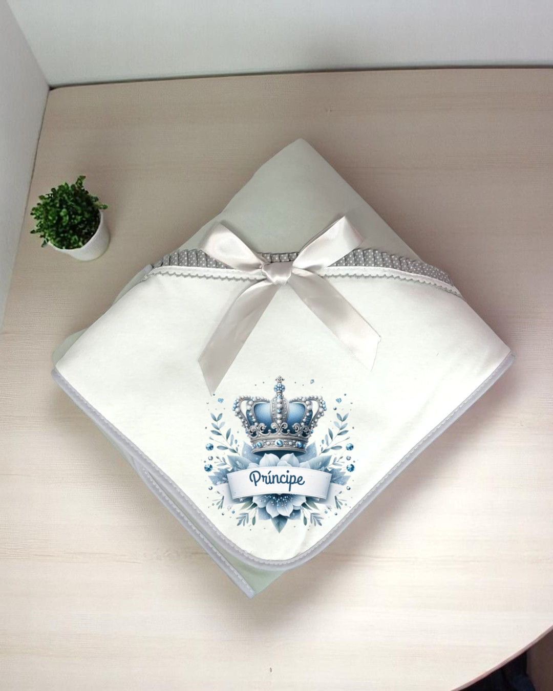 Manta branca com coroa azul e texto Príncipe em superfície de madeira com planta ao lado