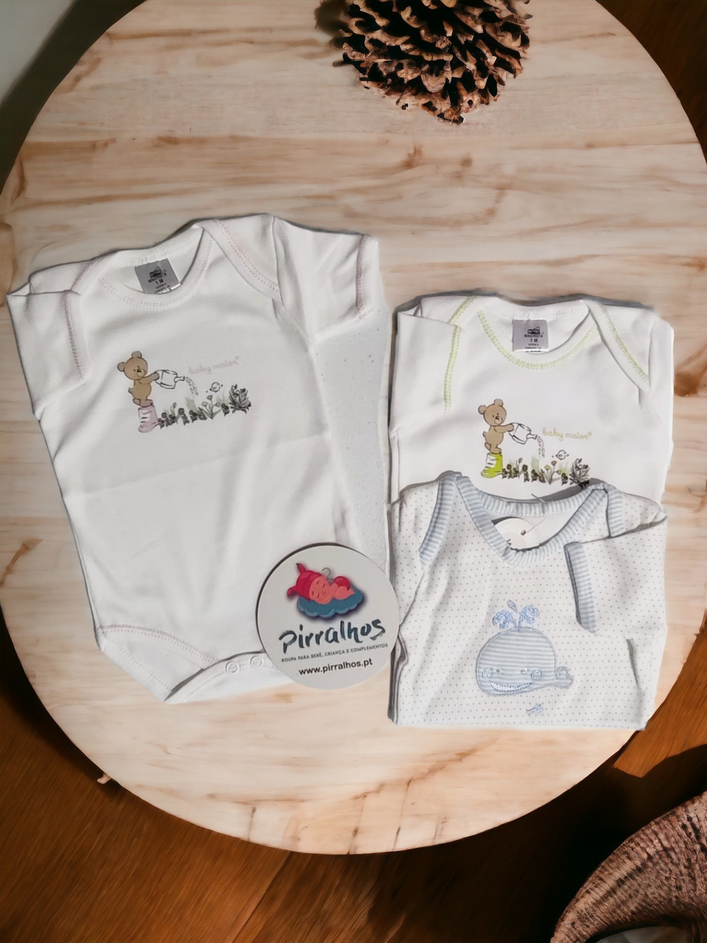 Três bodies de bebé com desenhos de animais, dois brancos e um cinza com padrão de pontos, sobre mesa de madeira clara