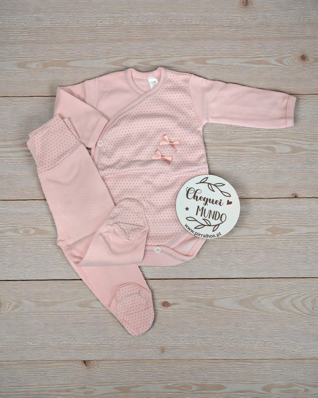 Conjunto de bebé cor-de-rosa com detalhes e placa CHEGUEI MUNDO sobre madeira clara.