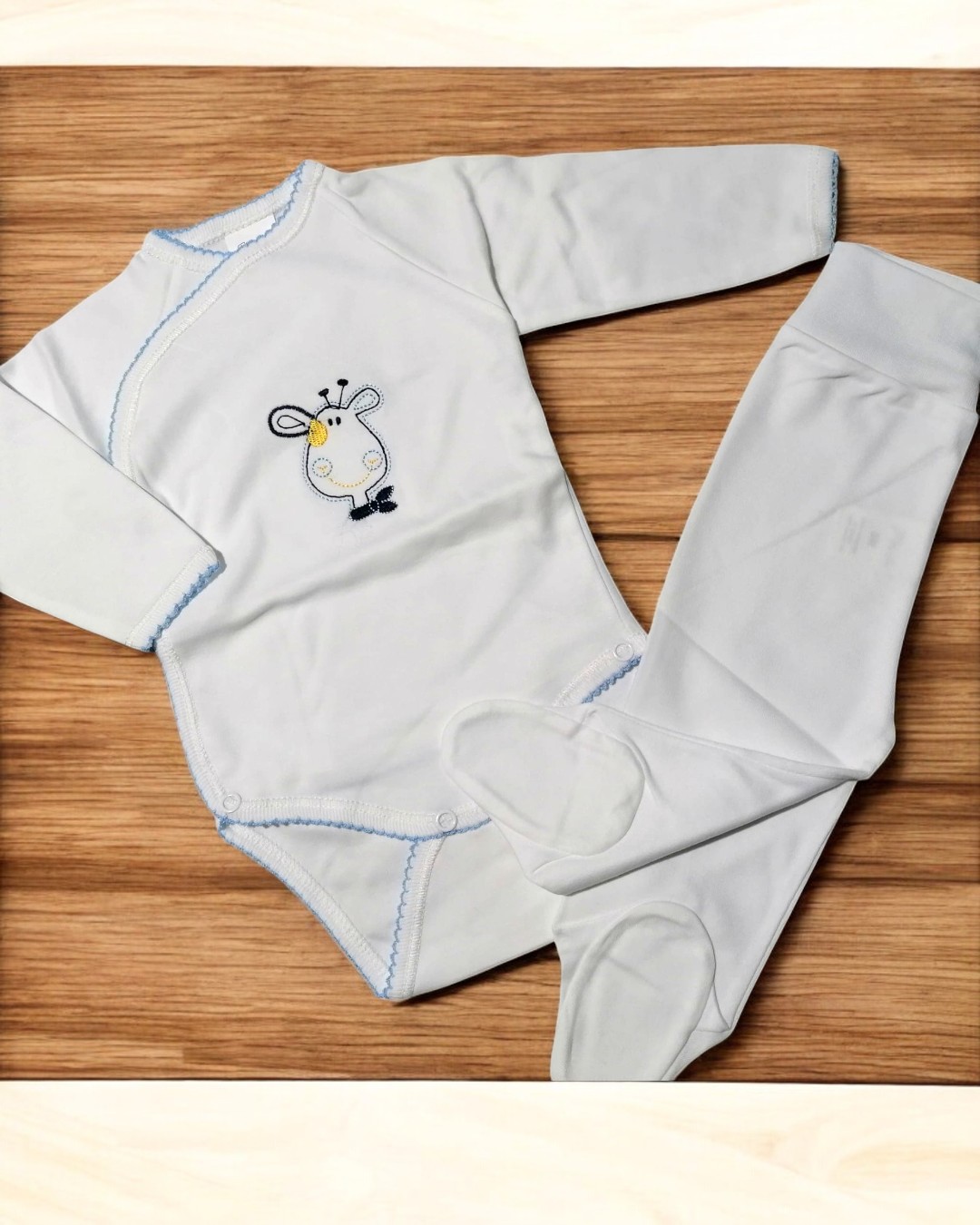 Conjunto de bebé branco com bordado de girafa e calças com pezinhos sobre madeira clara