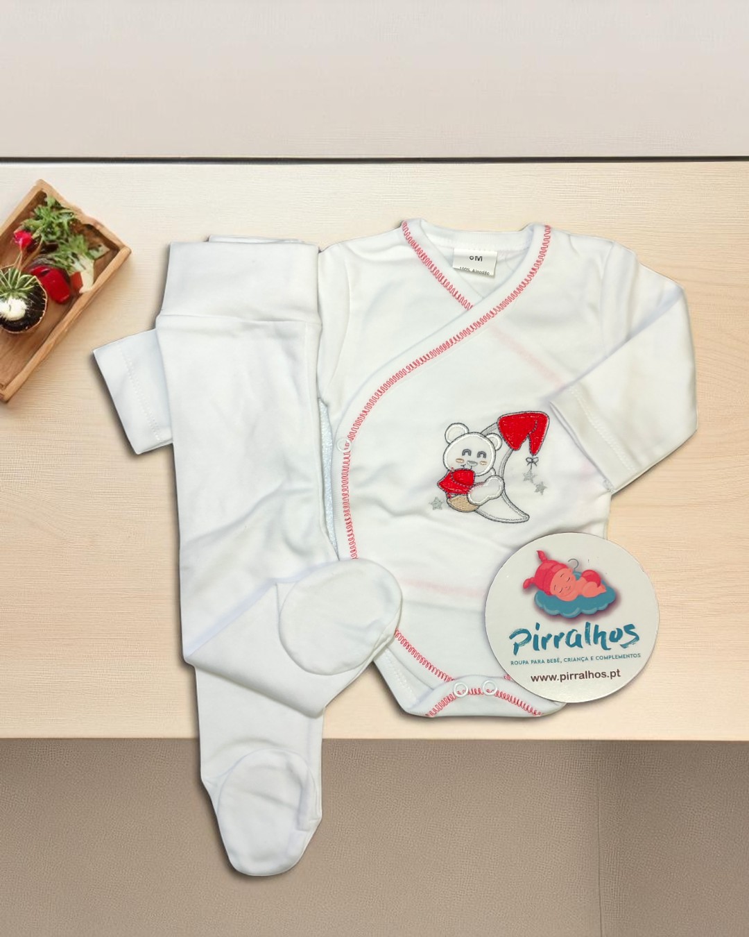 Conjunto de bebé branco com bordado de ursinho e gorro vermelho, acompanhado de etiqueta Pirralhos e vaso de plantas