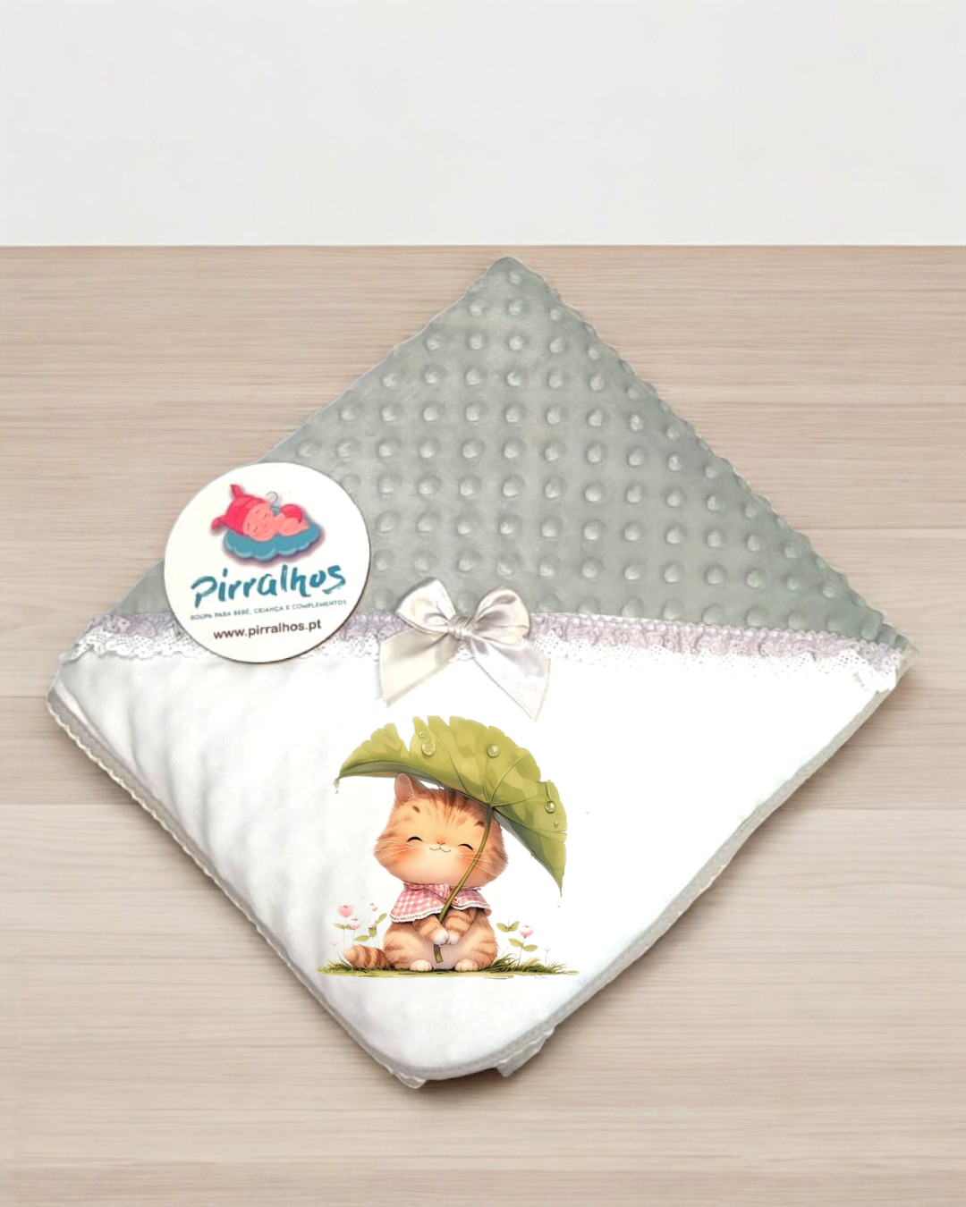 Manta de bebé branca e cinza com desenho de gatinho e laço, etiqueta Pirralhos, sobre superfície de madeira.