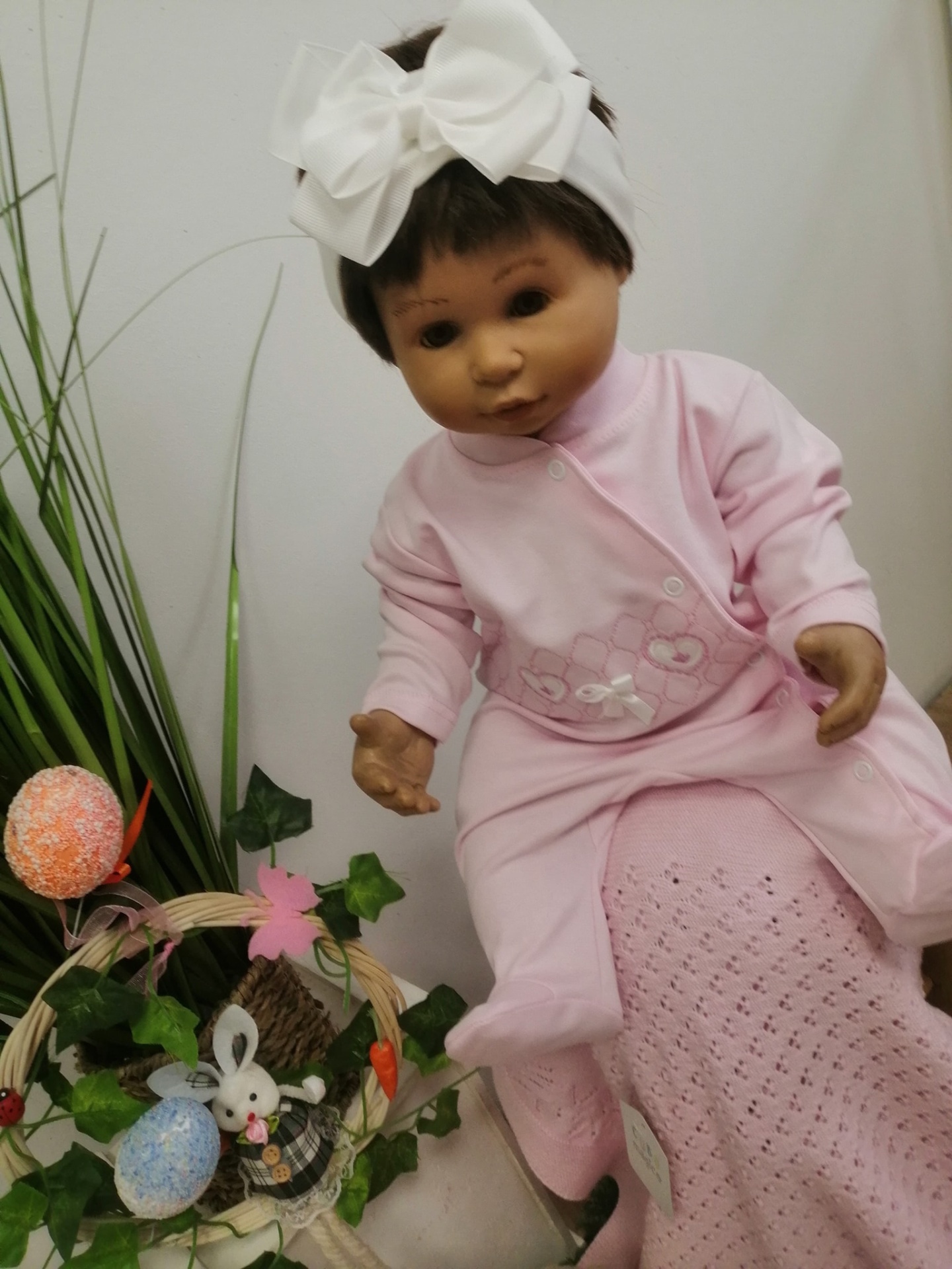 Boneca bebé com roupa rosa e laço branco, junto a cesto decorativo com ovos e coelho