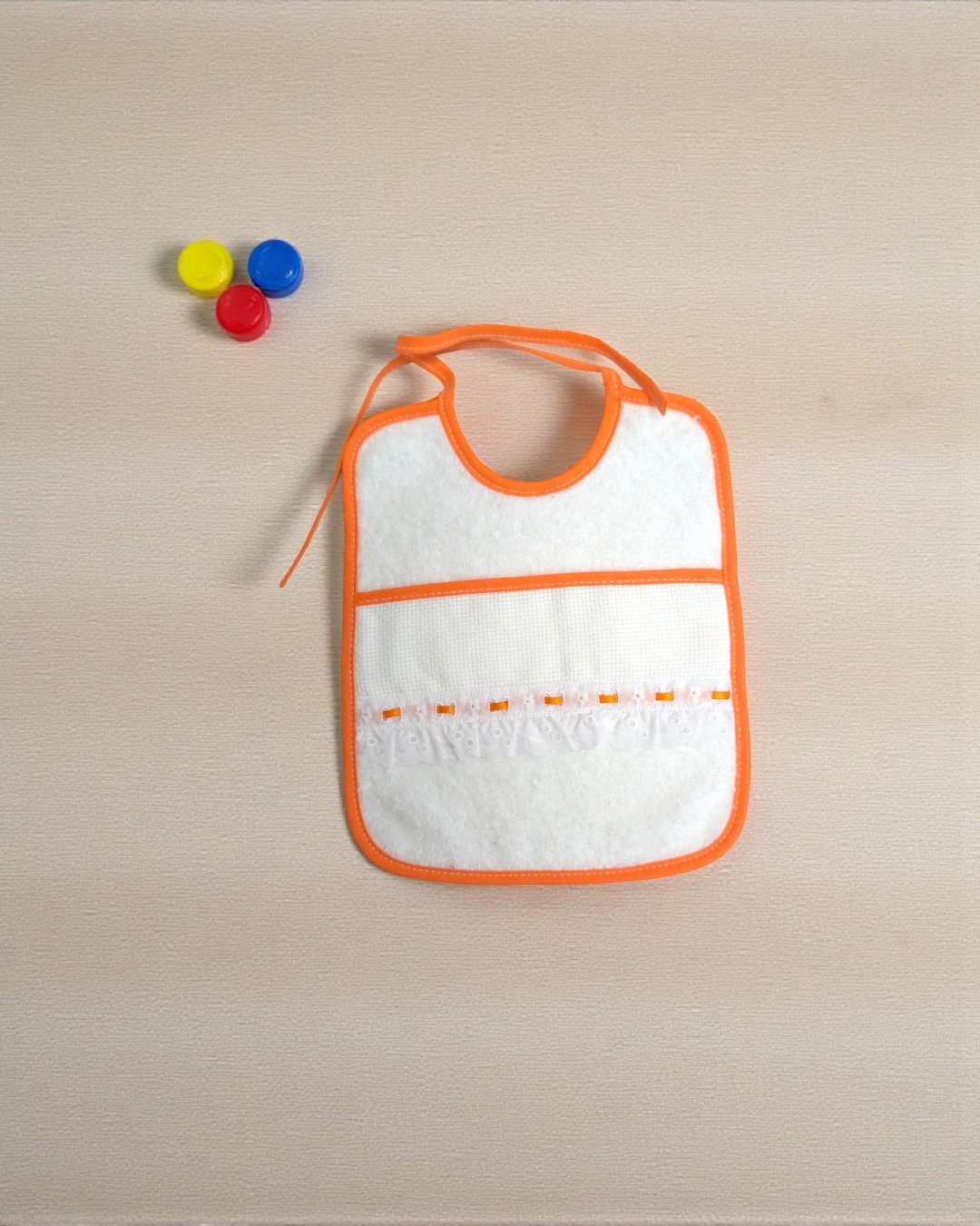 Babete branco com bordas laranja e fita decorativa, ao lado tampas amarelo, azul e vermelho