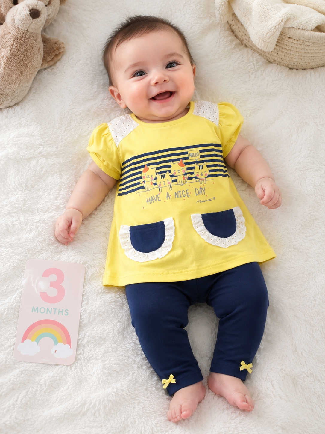 Bebé com túnica amarela e leggings azuis deitado em tecido branco, a sorrir, com cartão rosa com 3 MONTHS e arco-íris ao lado.