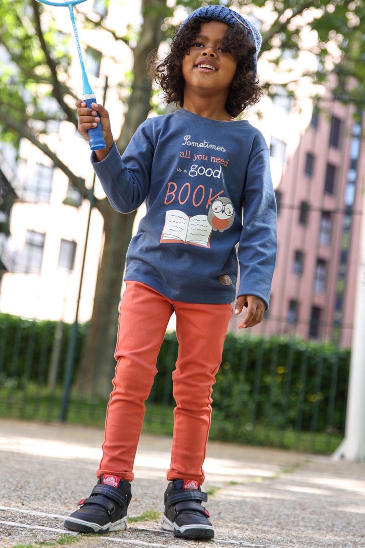 Criança com camisola azul com texto e ilustração, calças laranja, sapatos pretos e gorro azul no exterior