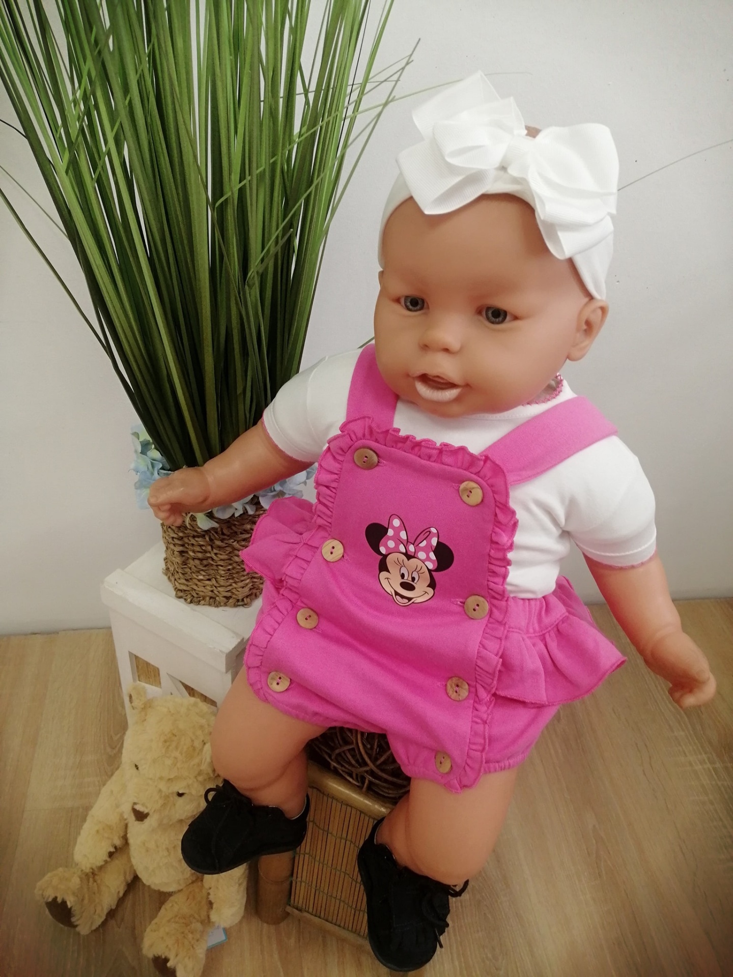 Boneca bebé com roupa rosa e branca, fita branca na cabeça, junto a urso de peluche e planta verde