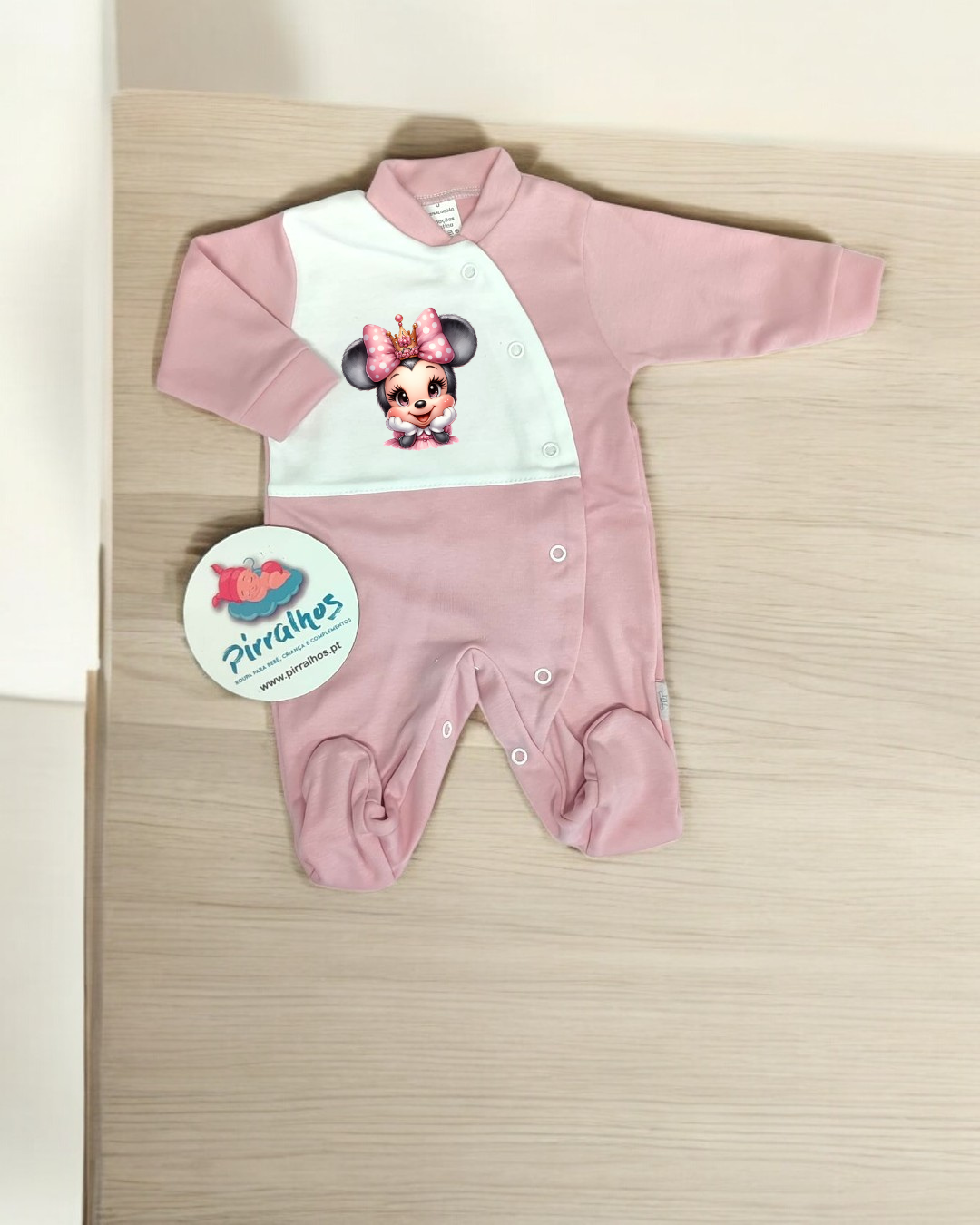 Macacão de bebé rosa e branco com estampa Minnie Mouse e botões, sobre superfície de madeira clara