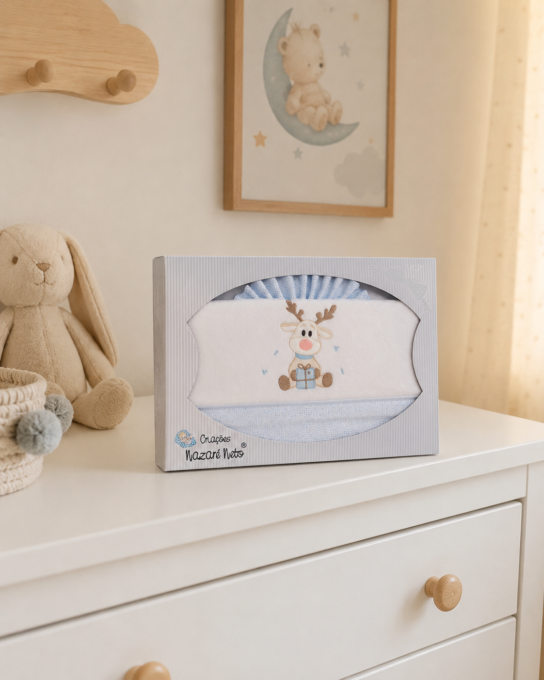 Caixa com toalha infantil bordada com rena a segurar presente numa cómoda branca com coelho de peluche ao lado