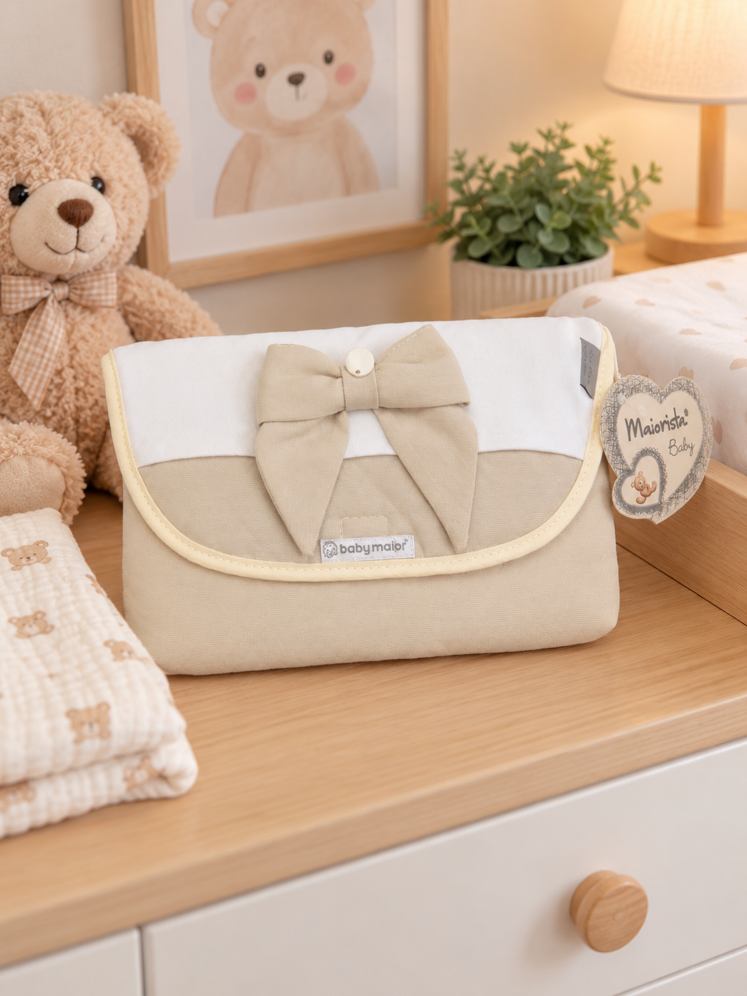 Necessaire de bebé bege e branca com laço, etiquetada baby major numa cómoda com ursinho de peluche e planta.