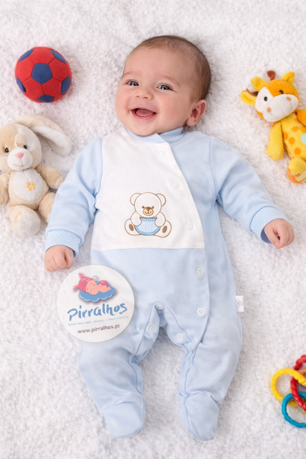 Bebé sorridente deitado numa manta branca com roupa azul clara com ursinho bordado e brinquedos ao redor