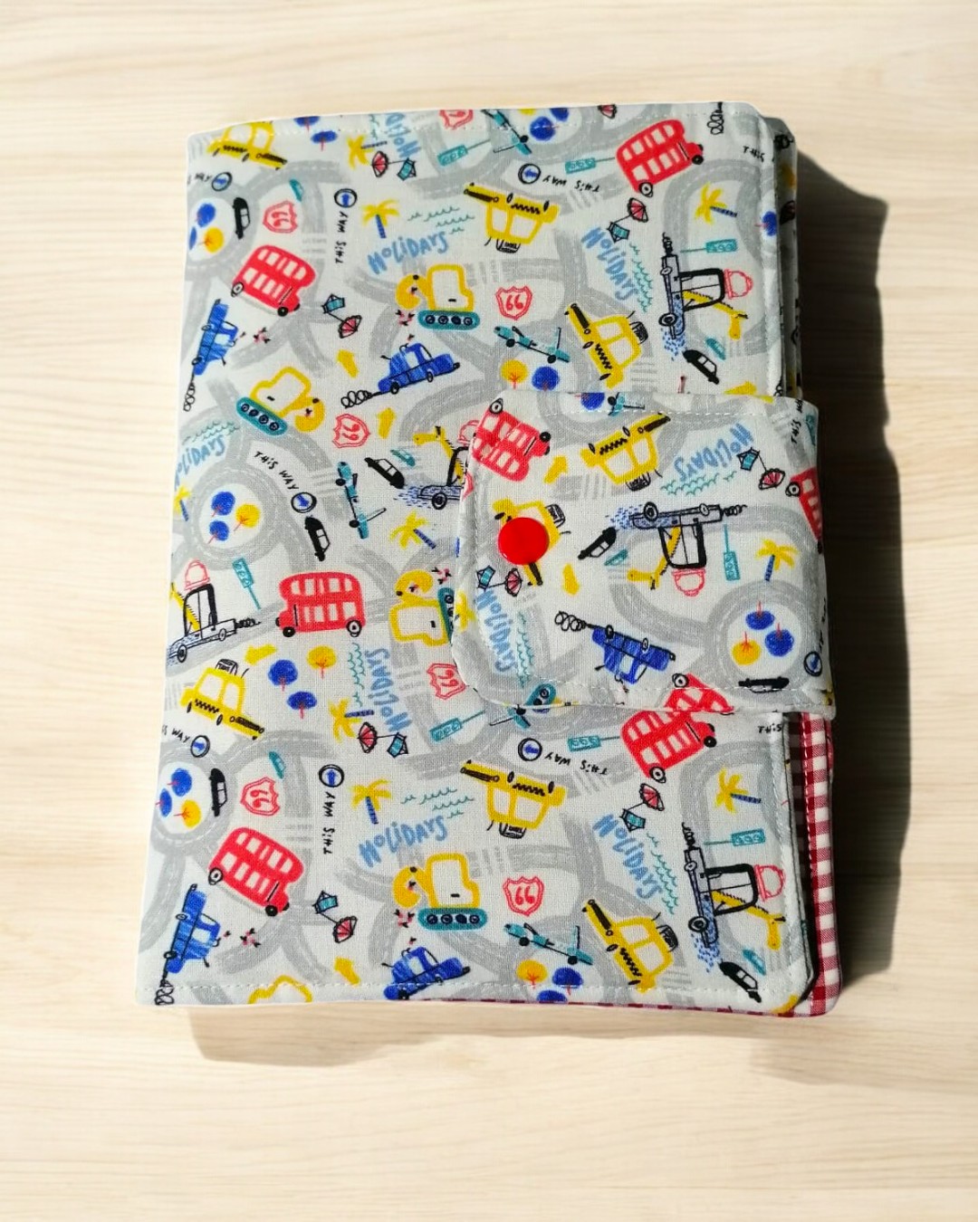 Produto de tecido com padrão colorido de veículos e texto, fecho botão vermelho, interior xadrez vermelho e branco.