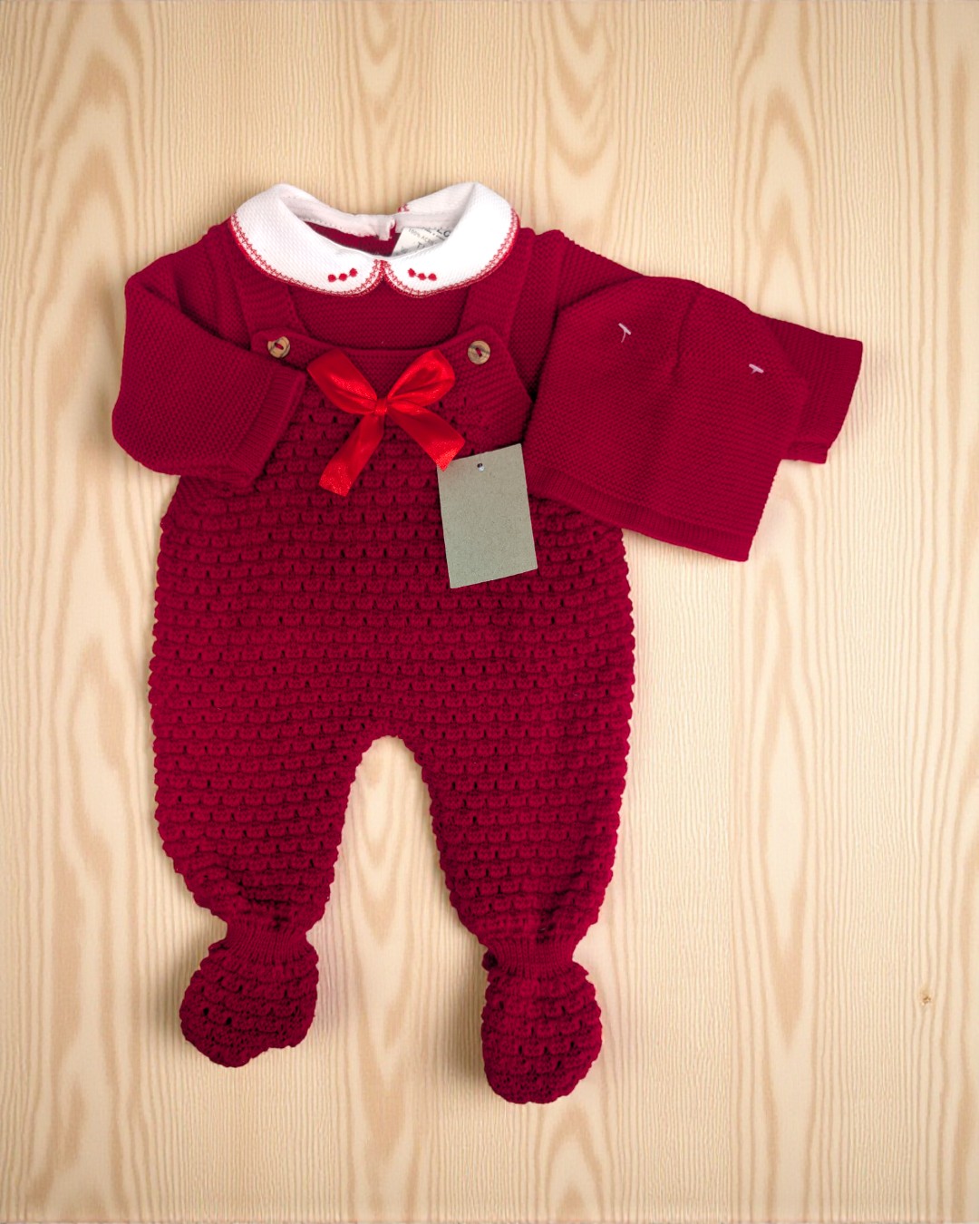 Conjunto de roupa para bebé vermelho escuro com macacão e gorro sobre fundo madeira clara