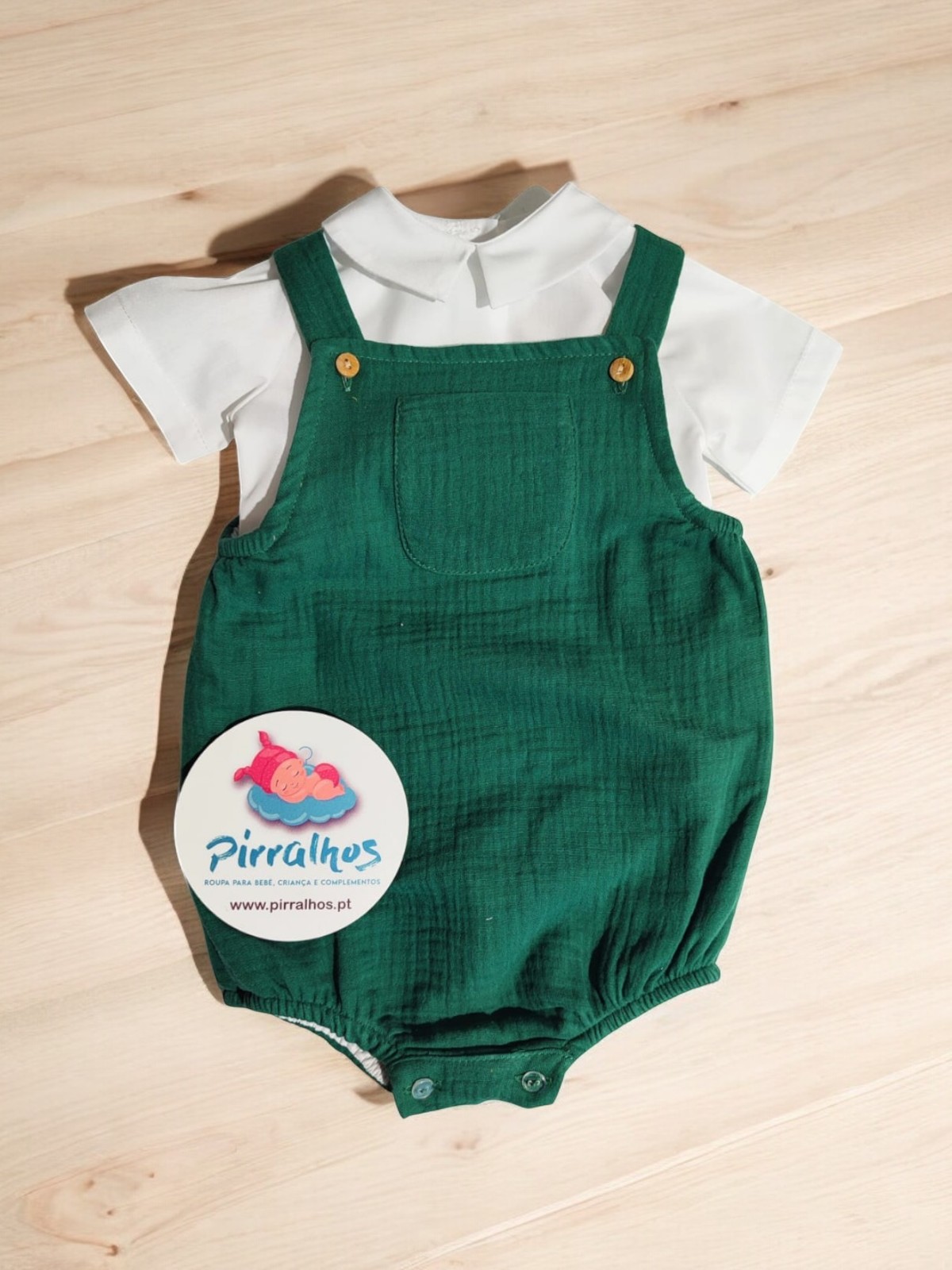 Conjunto de roupa de bebé com body branco e jardineira verde sobre superfície de madeira clara