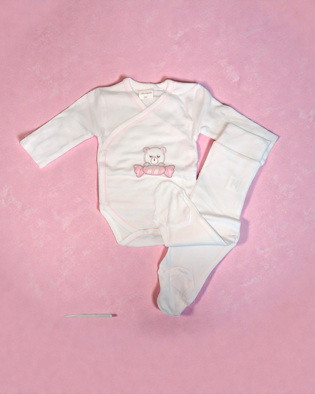 Conjunto branco de roupa de bebé com bordado de urso e doce em fundo rosa.