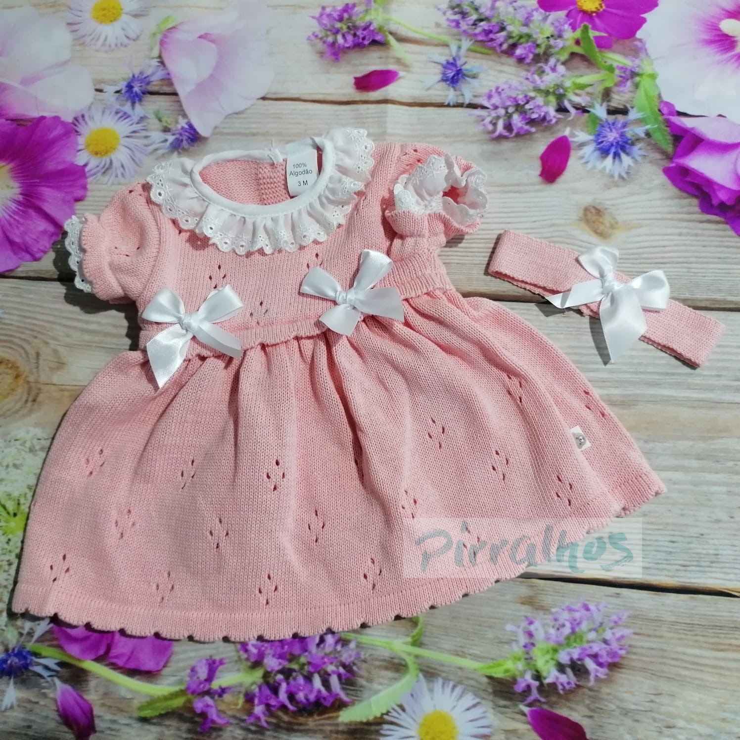 Vestido de bebé cor-de-rosa com gola branca e laços, com faixa de cabelo correspondente sobre superfície de madeira e flores