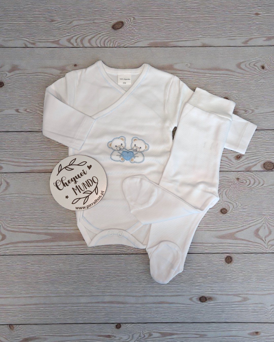 Conjunto bebé branco com bordado de ursos e placa redonda com texto em fundo de madeira.