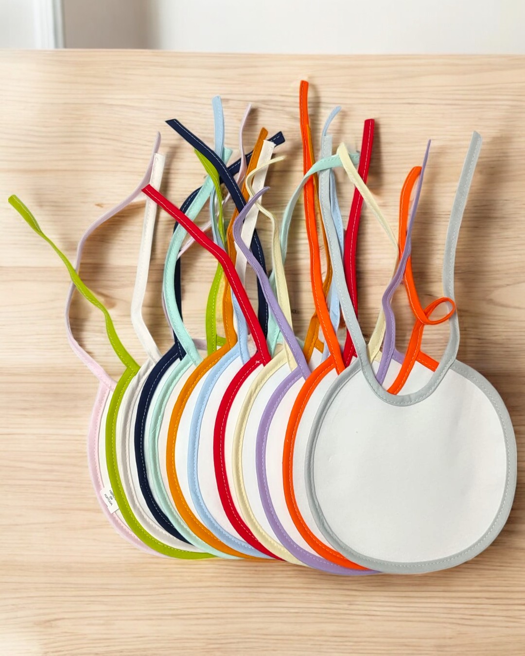 Babetes redondos coloridos para bebé com fitas para amarrar sobre madeira clara