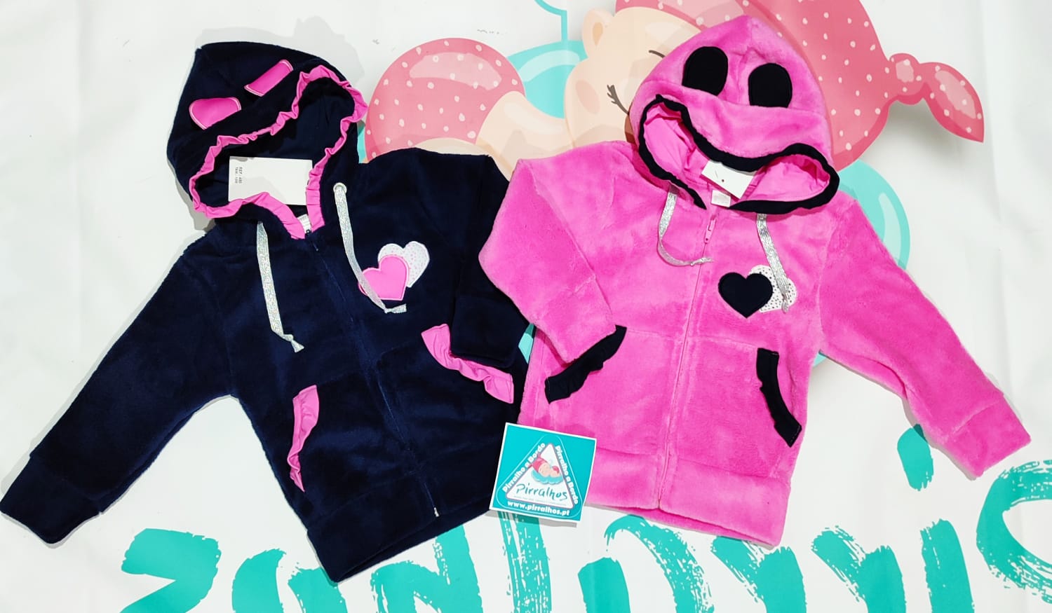 Casacas de bebé felpudas, azul escuro com detalhes cor-de-rosa e rosa com capuz decorado, sobre superfície branca colorida