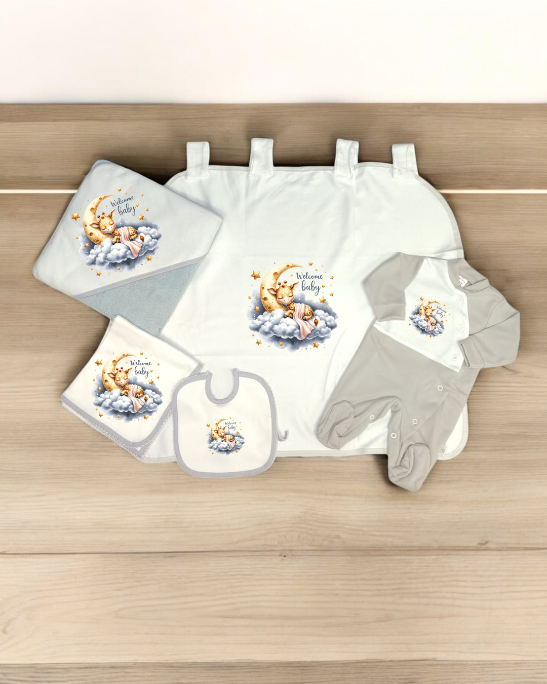 Conjunto de roupa de bebé e acessórios com estampado de ursinho e texto 'Welcome baby' em fundo cinzento e branco.