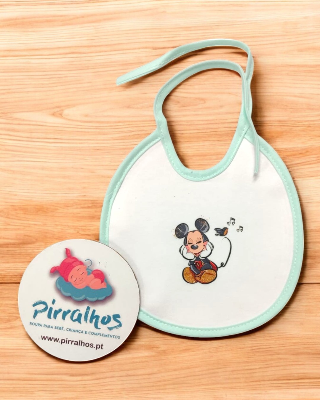 Babete branco com borda verde e estampa de Mickey Mouse ao lado de cartão redondo com logo e texto Pirralhos.