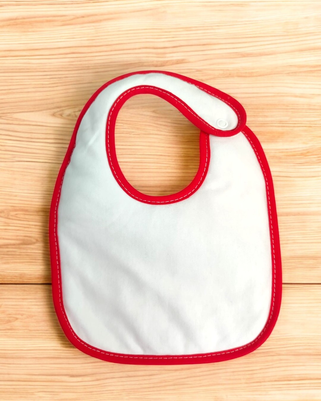 Babete branco com borda vermelha e fecho de botão de pressão sobre superfície de madeira