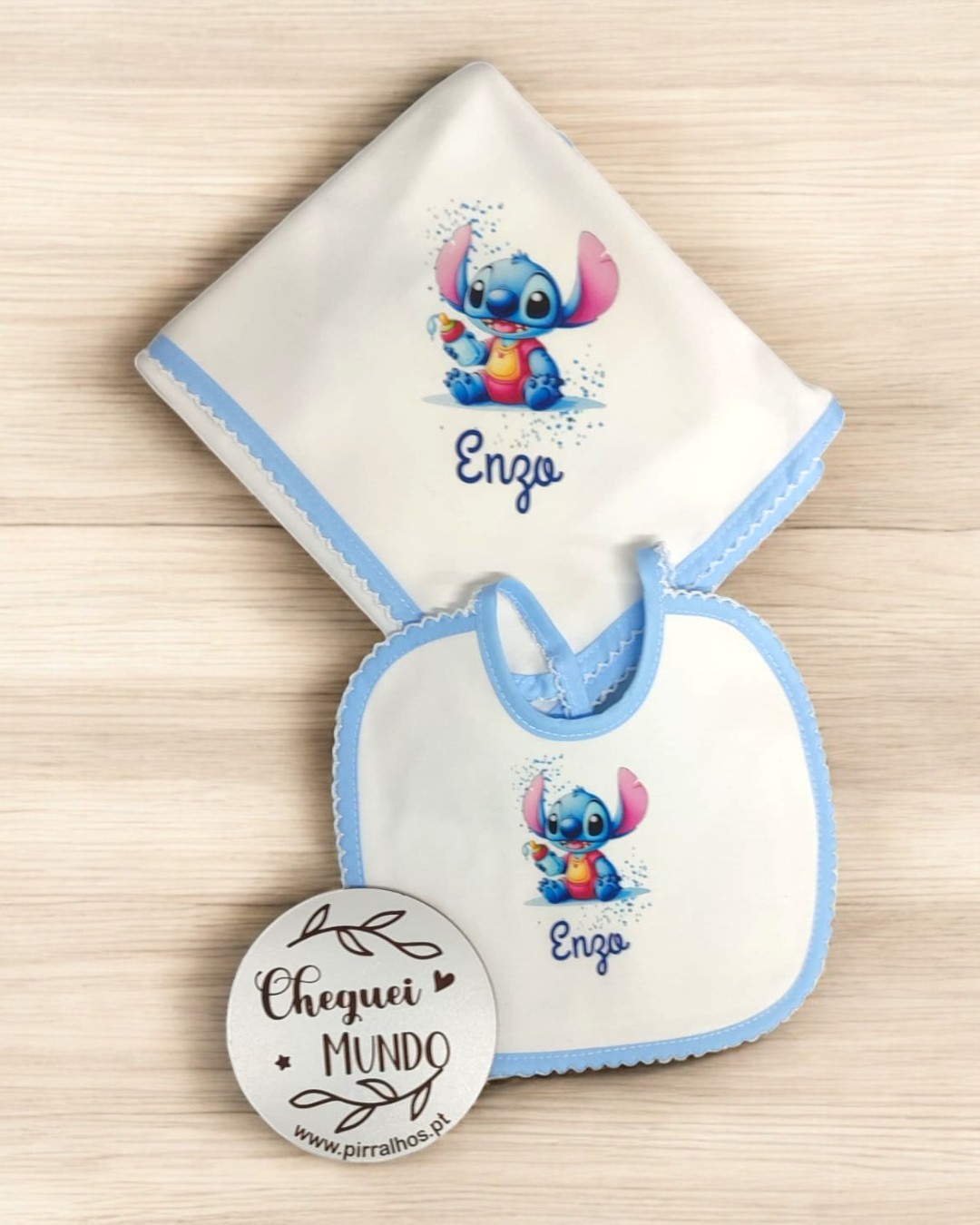 Babete e fralda de bebé com desenho de personagem azul e rosa e texto 'Enzo', sobre mesa clara com placa 'Cheguei MUNDO'.