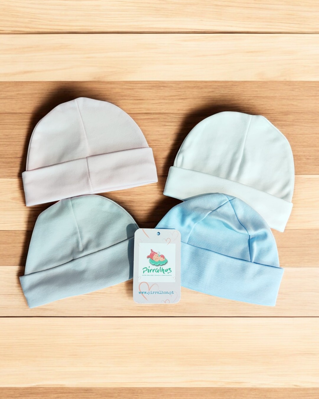 Quatro gorros de bebé em tecidos macios coloridos em tons pastel sobre uma mesa de madeira clara