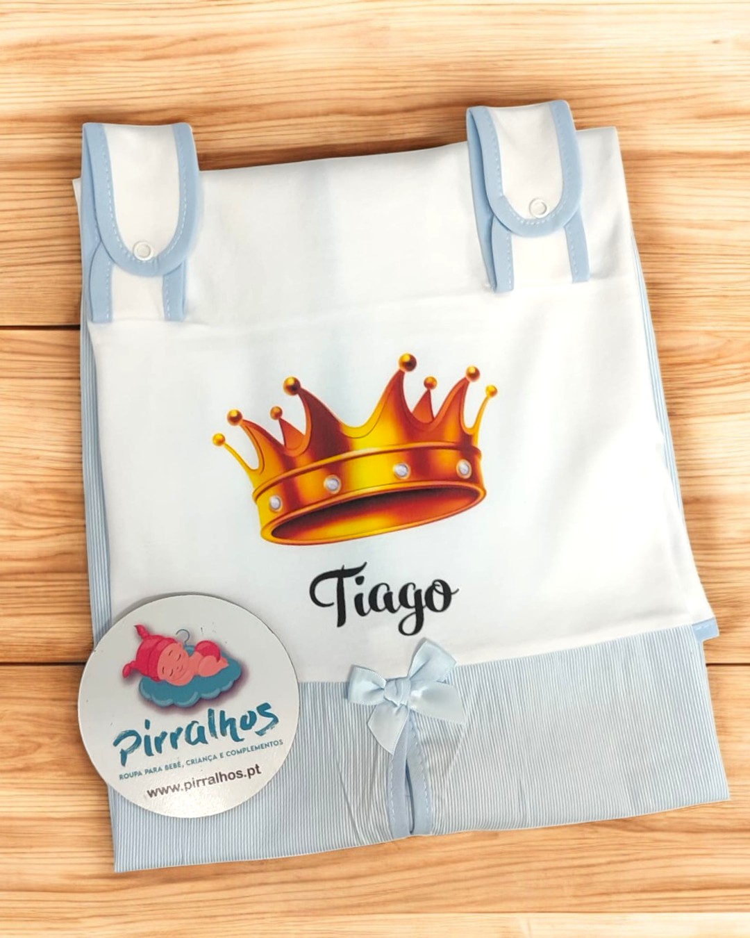 Capa para bebé branca com coroa amarela e o nome Tiago, detalhes em azul claro, sobre superfície de madeira