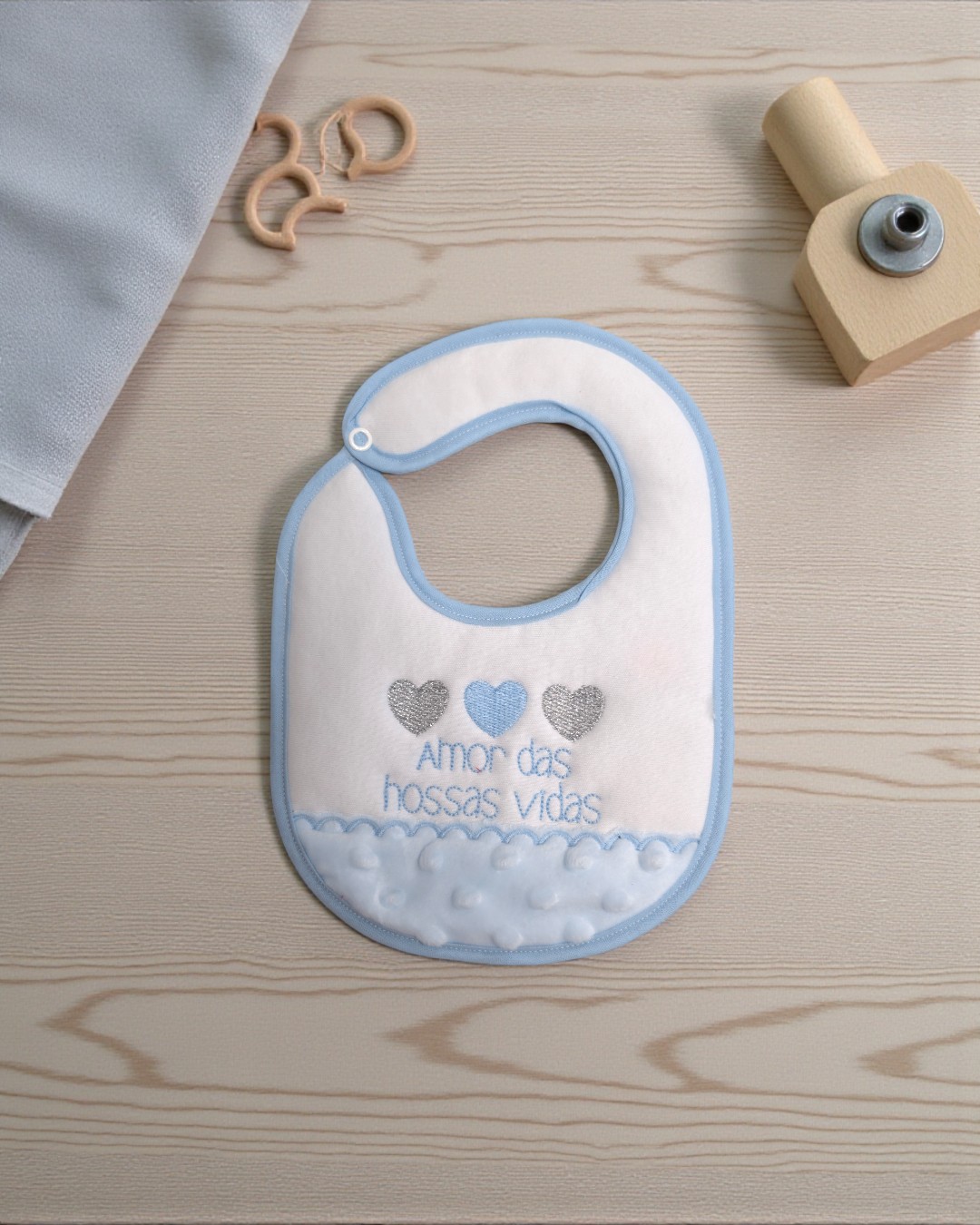 Babete de bebé branco com bordas azuis e texto bordado colocado sobre mesa de madeira clara.