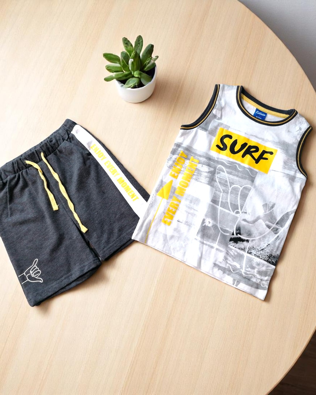 Conjunto infantil de t-shirt sem mangas branca e calções cinza escuro sobre mesa clara com vaso de planta suculenta.