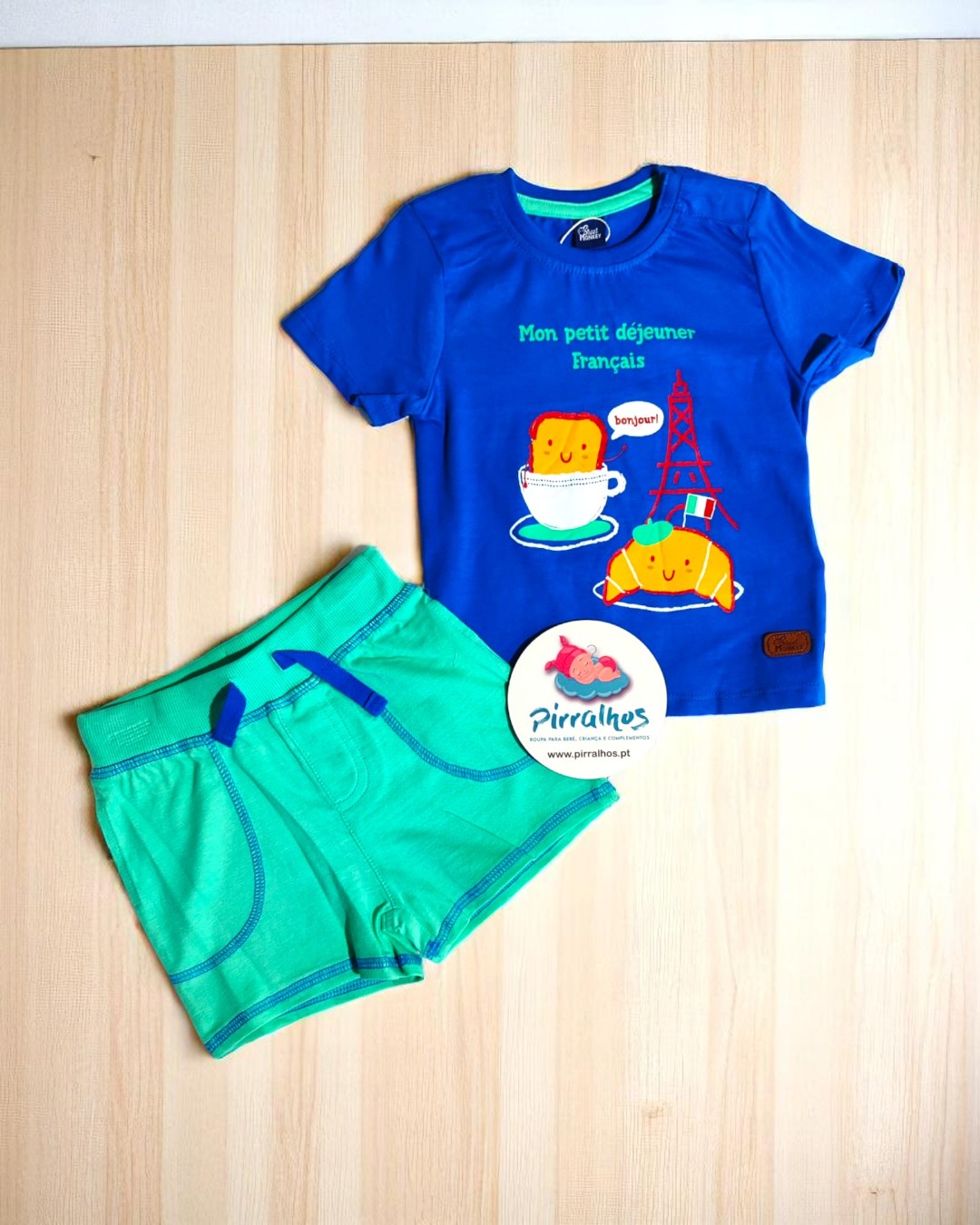 Conjunto infantil azul e verde com texto e estampa colorida sobre fundo de madeira clara.