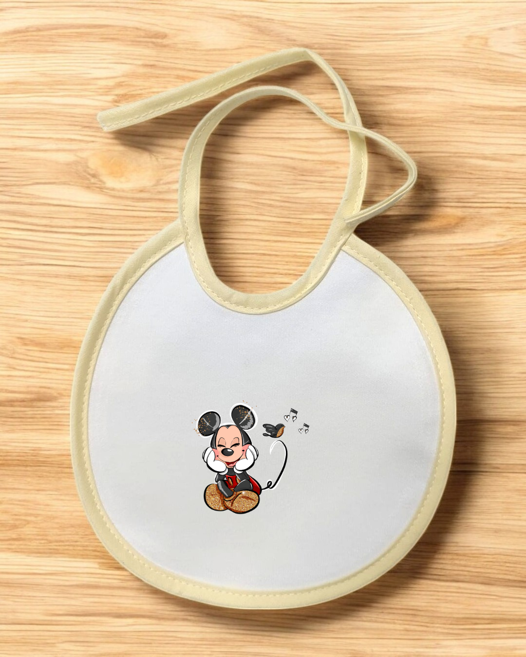 Babete de bebé branco com bordas bege e desenho de Mickey Mouse musical sobre superfície de madeira.